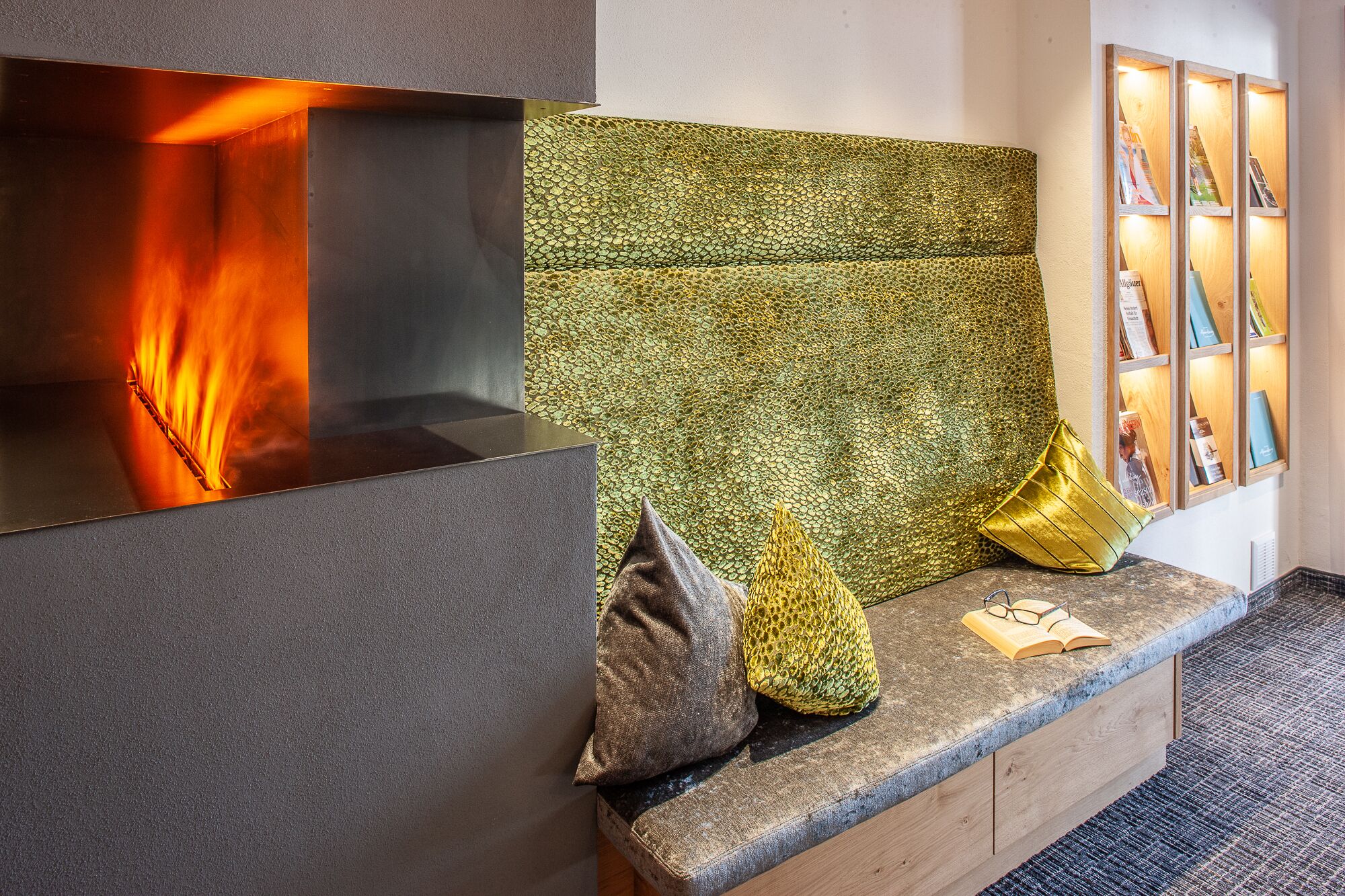 A modern fireplace with a textured green wall behind it, a bench with cushions, and a display of framed pictures on the wall.