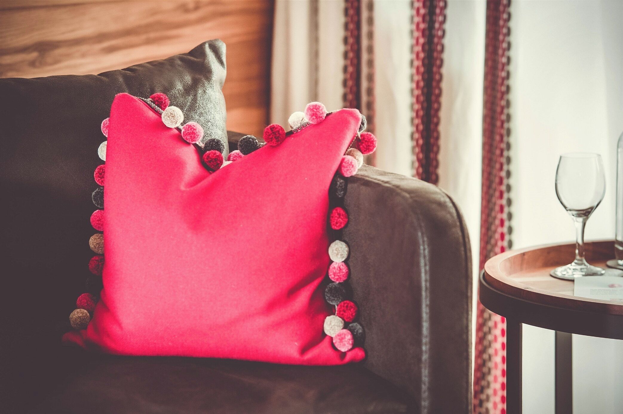 A red pillow with pom-poms on a brown leather sofa.