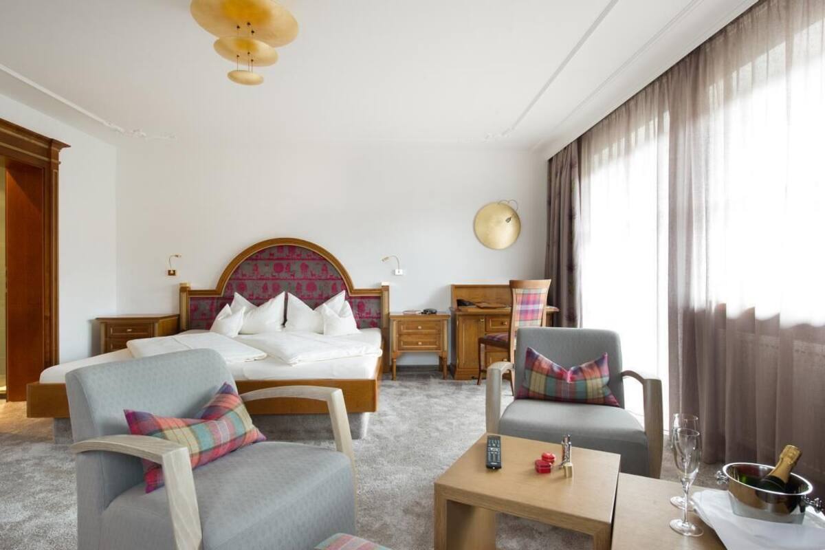 Une chambre d’hôtel comprenant un lit, deux fauteuils, une table de chevet, un bureau, une lampe, une horloge et une fenêtre avec des rideaux.