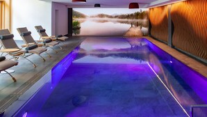 A modern indoor pool with a view of a lake and mountains, equipped with lounge chairs and a wooden deck. - Hotel Alpenkönig (Oberstaufen)