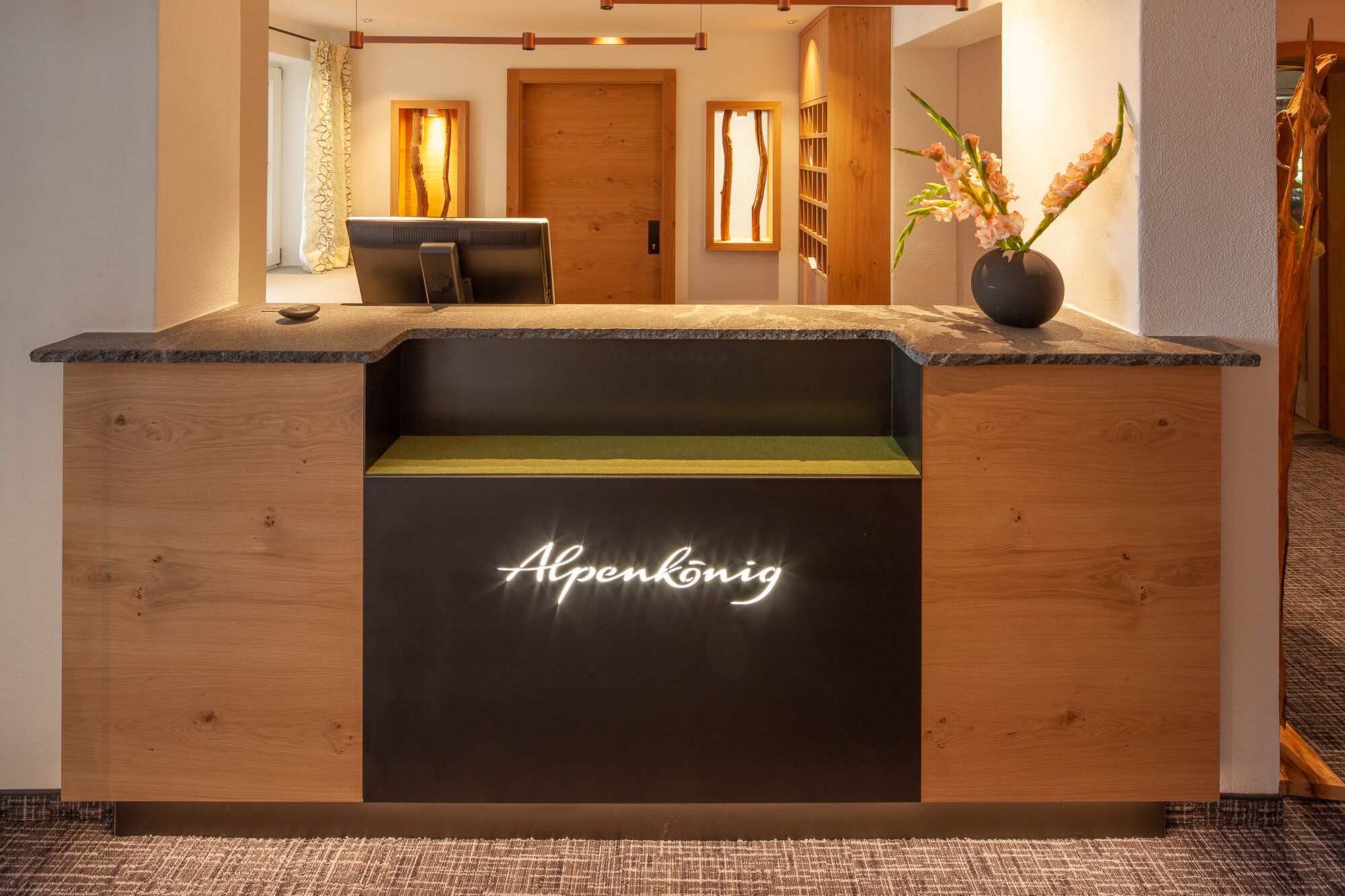 Un mostrador de recepción con detalles de madera, un letrero de neón que dice "Alpenkönig" y un florero con flores en la encimera.