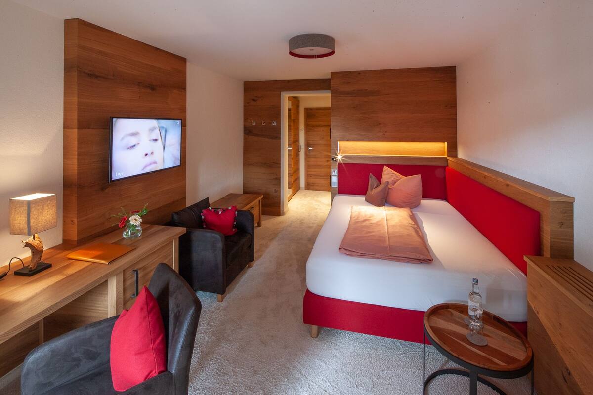 Une chambre d’hôtel avec un mur en bois, un lit avec une tête de lit rouge, un téléviseur fixé au mur, un bureau avec une lampe et une petite table avec une bouteille.