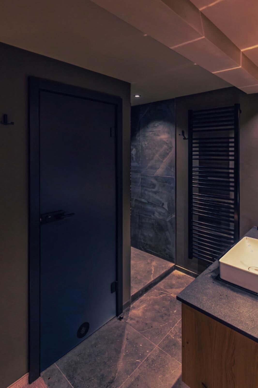 A hallway with a dark door, a tiled floor, and a bathroom with a shower and a towel rack.
