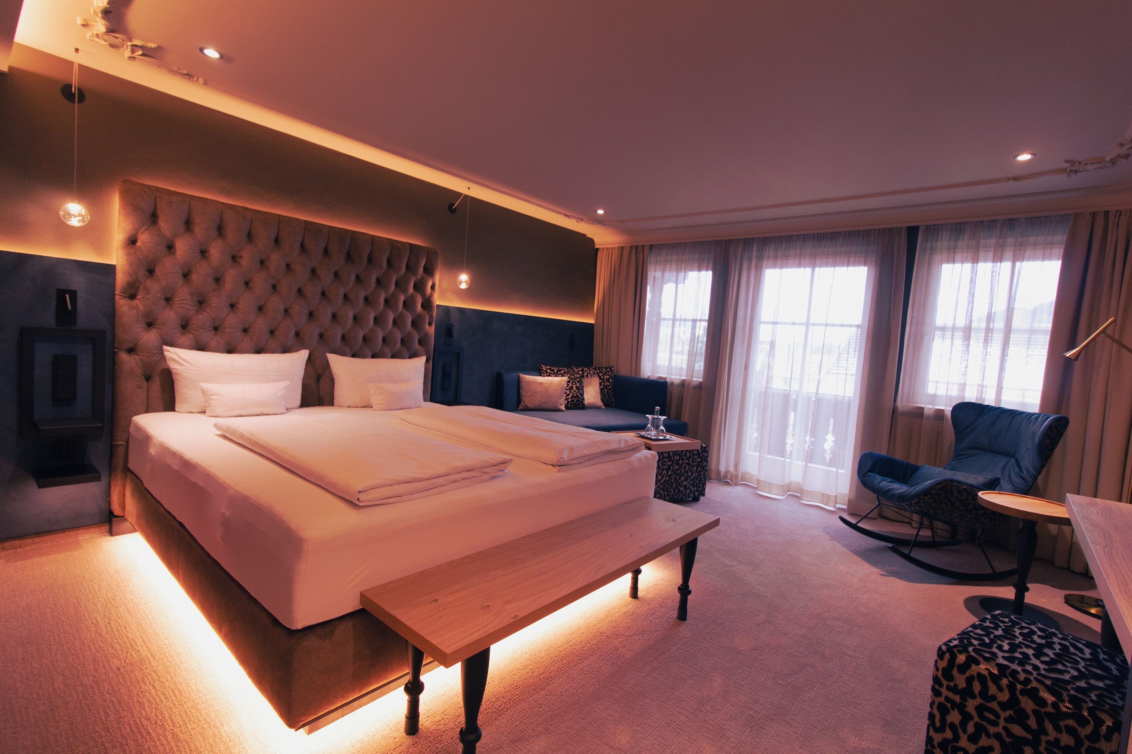 A modern hotel room with a large bed, a bench, a sofa, and a chair.