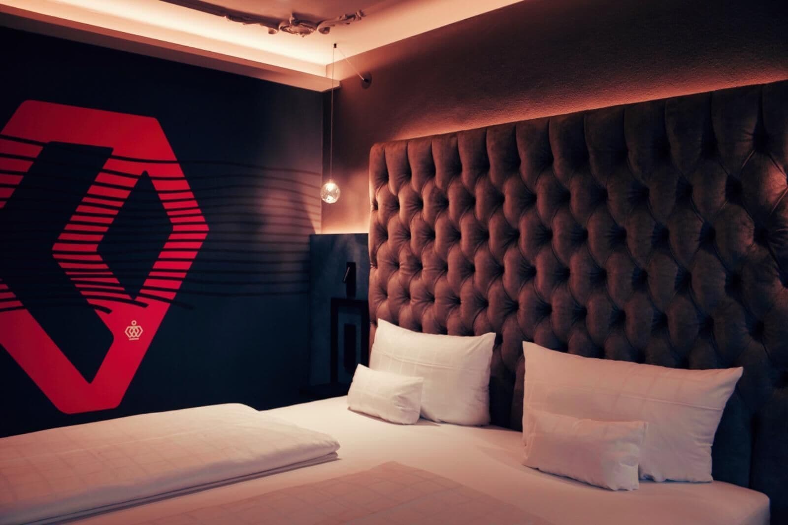 A modern hotel room with a tufted headboard, a large geometric logo on the wall, and a neatly made bed with white linens.