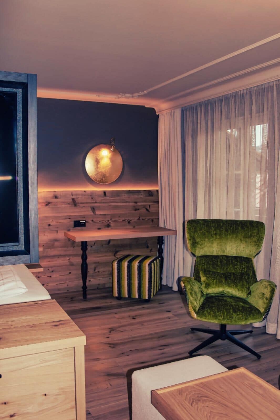 A room with a wooden wall, a round wall lamp, a green armchair, a wooden table, and a striped suitcase.