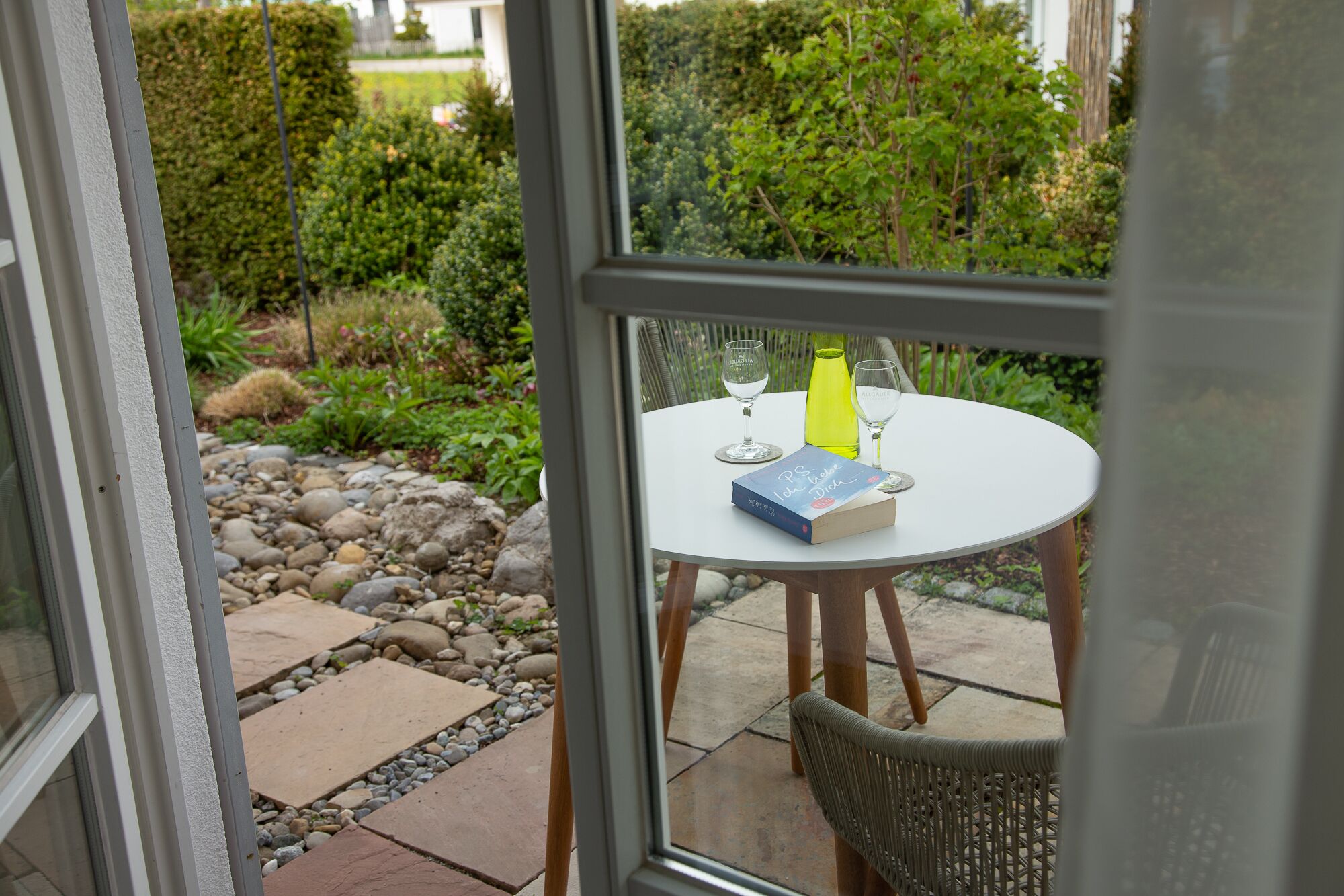 Eine Glastür führt zu einer Terrasse im Freien mit einem runden Tisch, zwei Weingläsern, einem Buch und einer gelben Flasche.