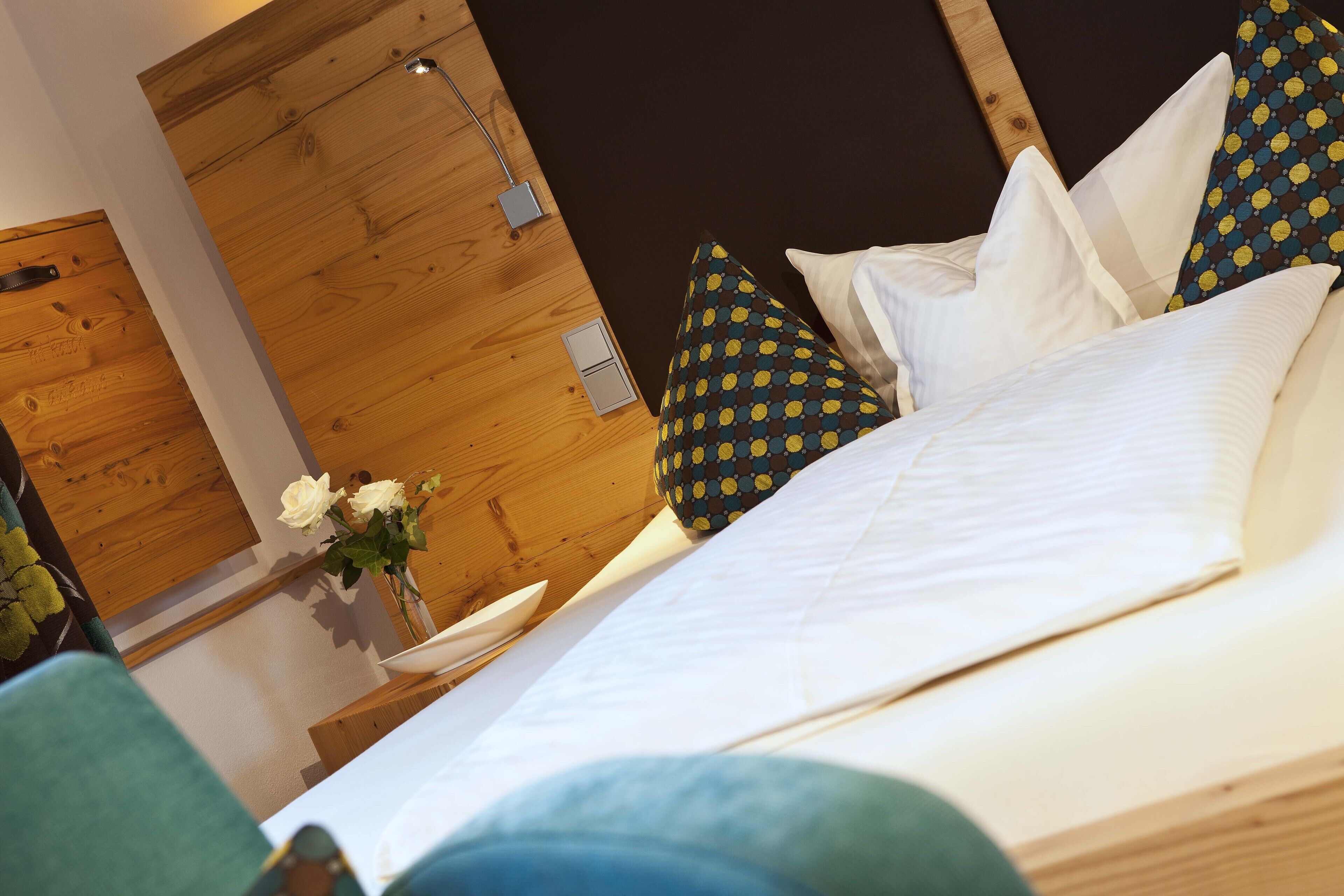 A neatly made bed with white linens and patterned pillows, a wooden headboard, and a small table with a vase of flowers.