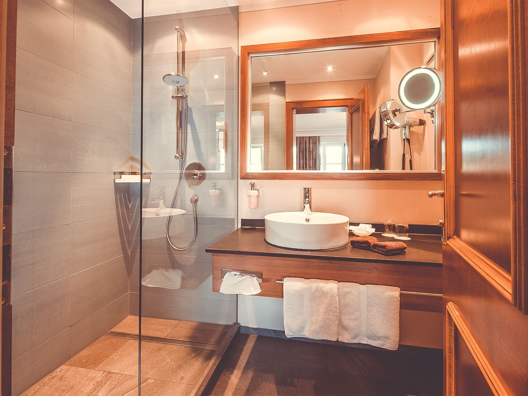 Un baño moderno con ducha envuelta en vidrio, tocador de madera, espejo grande y lavamanos.