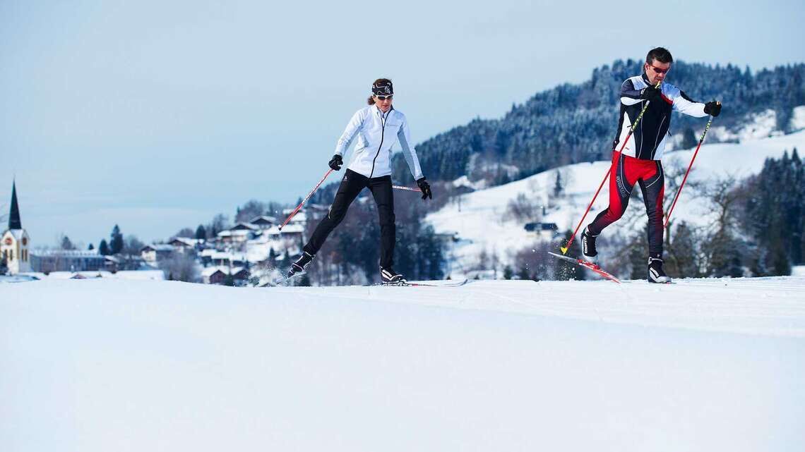 Deux personnes pratiquent le ski de fond dans un paysage enneigé, avec une église et des maisons en arrière-plan.