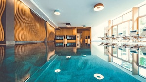 A long, reflective hallway with a pool, wooden wall panels, and a row of sun loungers by a large window. - Hotel Alpenkönig (Oberstaufen)