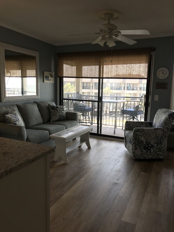 A living room with a gray sofa, a white coffee table, and a patterned armchair. A ceiling fan is visible, and there is a sliding glass door leading to an outdoor balcony.