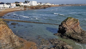 A coastal view with a rocky shoreline, clear blue water, and buildings in the background. - Nice chalet in a residence in St Hilaire (Saint-Hilaire-de-Riez)