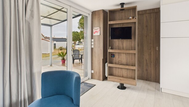 A modern room with a sliding glass door leading to a patio, a blue chair, a wooden shelving unit, and a flat-screen TV mounted on the wall. - Nice chalet in a residence in St Hilaire (Saint-Hilaire-de-Riez)