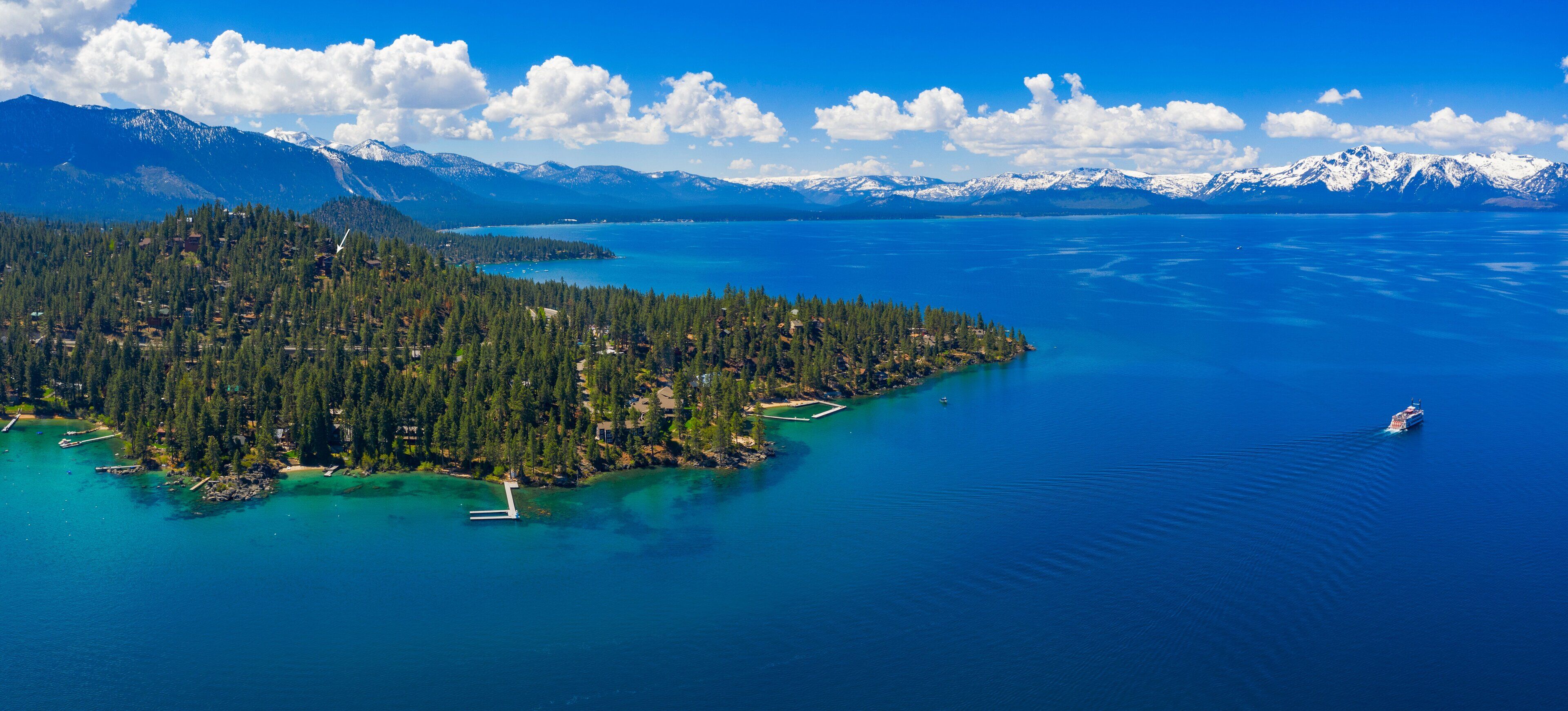 An aerial view of a coastal area with a forested island, clear blue waters, and distant snow-capped mountains.