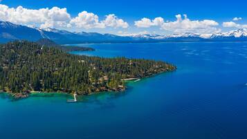 An aerial view of a coastal area with a forested island, clear blue waters, and distant snow-capped mountains.
