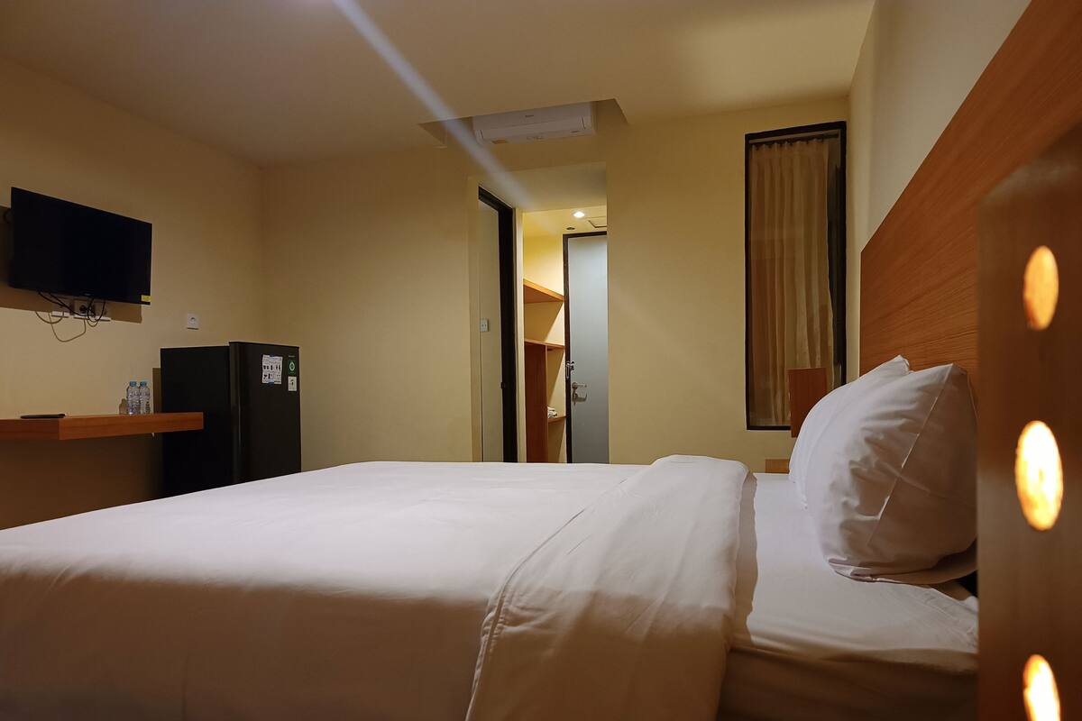 A hotel room with a bed, a television mounted on the wall, a refrigerator, and a wooden headboard.