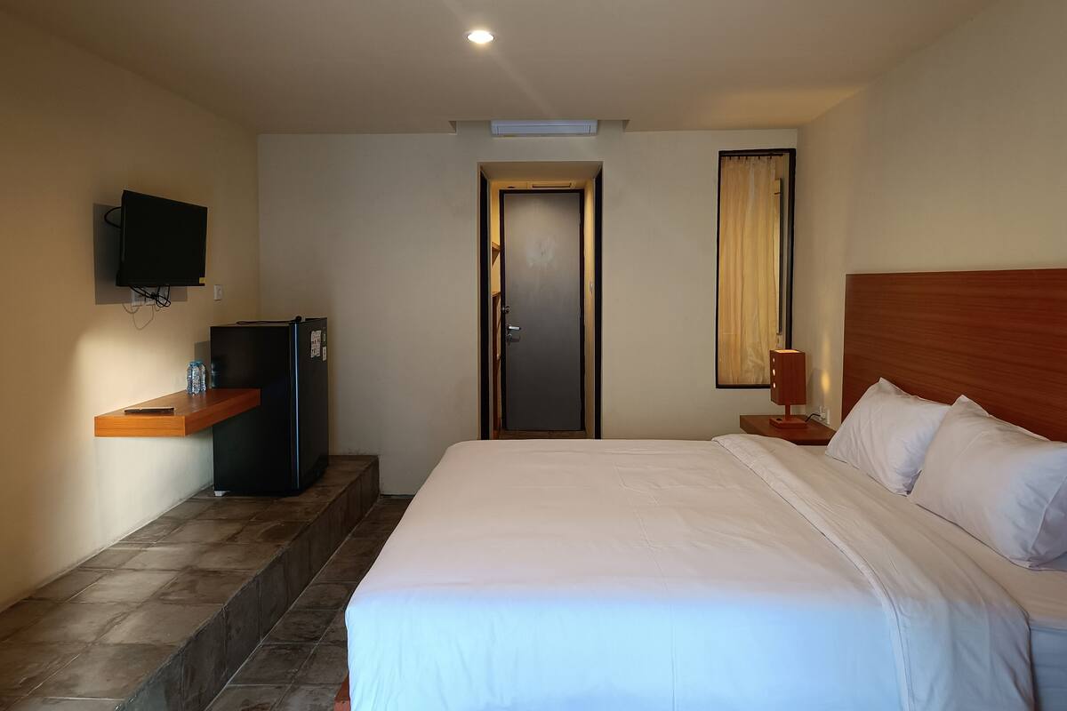 A hotel room with a bed, a television mounted on the wall, a refrigerator, a wooden nightstand, and a door.