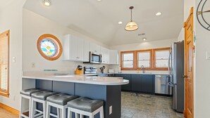 A kitchen with a central island, bar stools, a microwave, a refrigerator, and a window with blinds. - Sea-U-Later!: 820 Ft to Beach Access; Private Pool, Hot Tub (Duck)