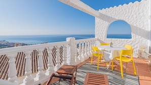 A terrace with a white balustrade, yellow chairs, a table, and a view of the sea.