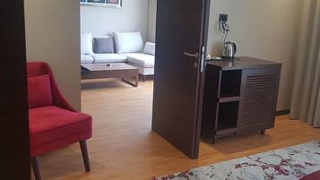 Habitación de hotel con piso de madera, un sillón rojo, un sofá, un mueble bajo y una alfombra estampada.