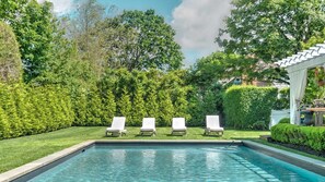 Ein rechteckiger Pool mit vier weißen Liegestühlen, die entlang der langen Seite angeordnet sind, umgeben von gepflegten Hecken und Bäumen.