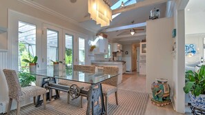 Una cocina amplia con una isla central, ventanas grandes y un comedor con mesa de vidrio y sillas de mimbre.