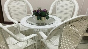 A round glass table surrounded by four white wicker chairs with a small vase of flowers on top. - Chogun Hostel (Chumphon)
