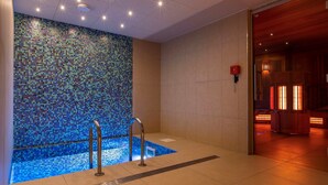 Un espace spa moderne agrémenté d’un mur en mosaïque, d’une petite piscine et d’un sauna visible à travers une porte ouverte.