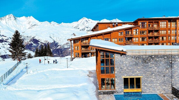 Une station de montagne enneigée, avec un bâtiment en bois doté d’une façade en pierre et d’un espace piscine.