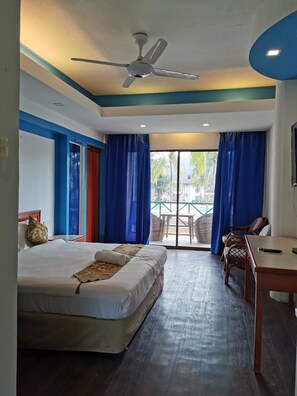 A hotel room with a bed, a desk, a chair, a ceiling fan, and a view of the outside.
