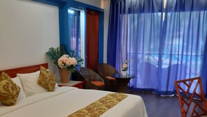 Een hotelkamer met een groot bed, een houten vloer, een plafondventilator, een raam met blauwe gordijnen en een tafeltje met een bloevas.