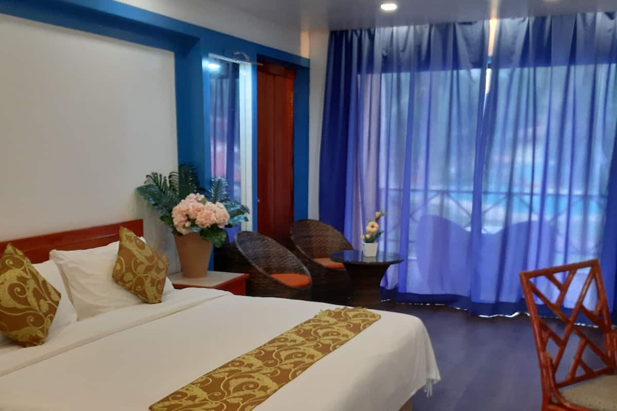 Een hotelkamer met een groot bed, een houten vloer, een plafondventilator, een raam met blauwe gordijnen en een tafeltje met een bloevas.