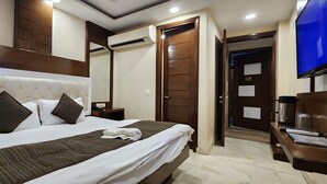 A hotel room with a large bed, a wooden headboard, a flat-screen TV, a bedside table with a lamp, and a door leading to another room. - TG Tashkent Hotel (New Delhi)