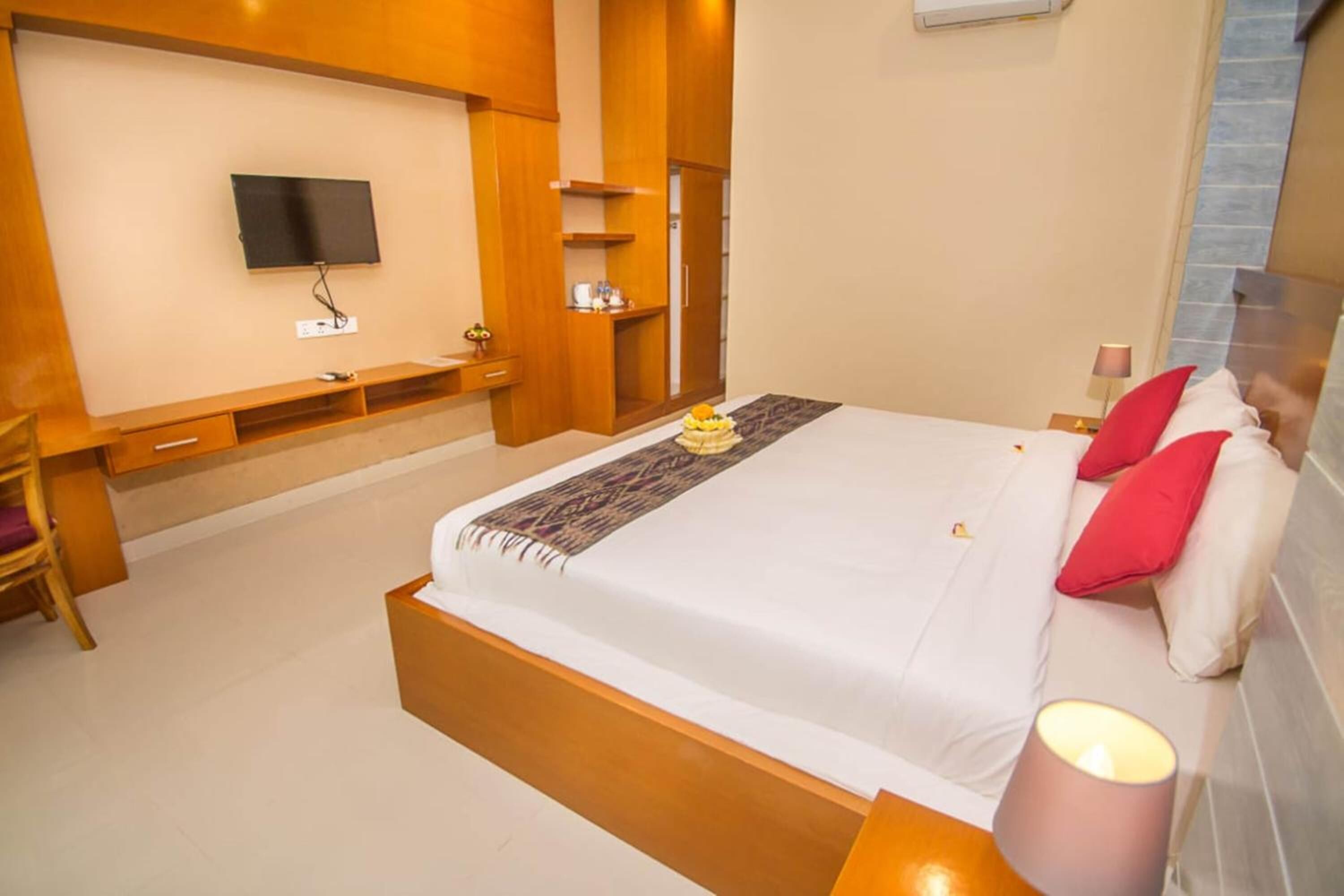 A modern hotel room with a large bed, a flat-screen TV mounted on the wall, a wooden bench, and a small table with a lamp.