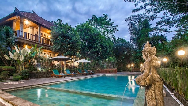 A swimming pool with lounge chairs and umbrellas, surrounded by lush greenery and a building with a balcony. - Adi Jaya Cottages Jungle Suites by EPS (Ubud)