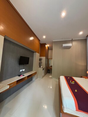 A hotel room with a bed, a TV mounted on the wall, a desk with a chair, and a small kitchenette area. - Adi Jaya Cottages Jungle Suites by EPS (Ubud)