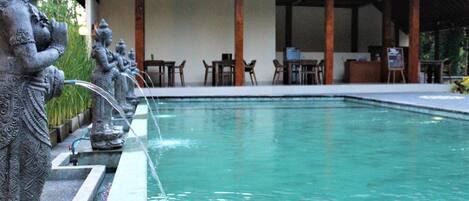 A pool with a statue on the left, a covered seating area with tables and chairs in the background.