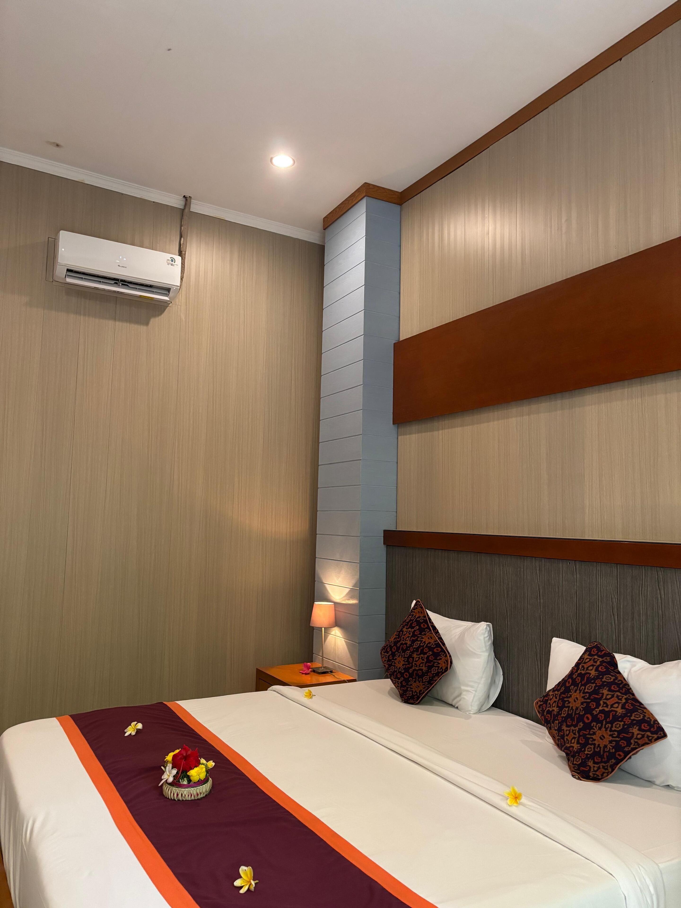 A hotel room with a bed, a nightstand, an air conditioning unit, and a wall-mounted lamp.