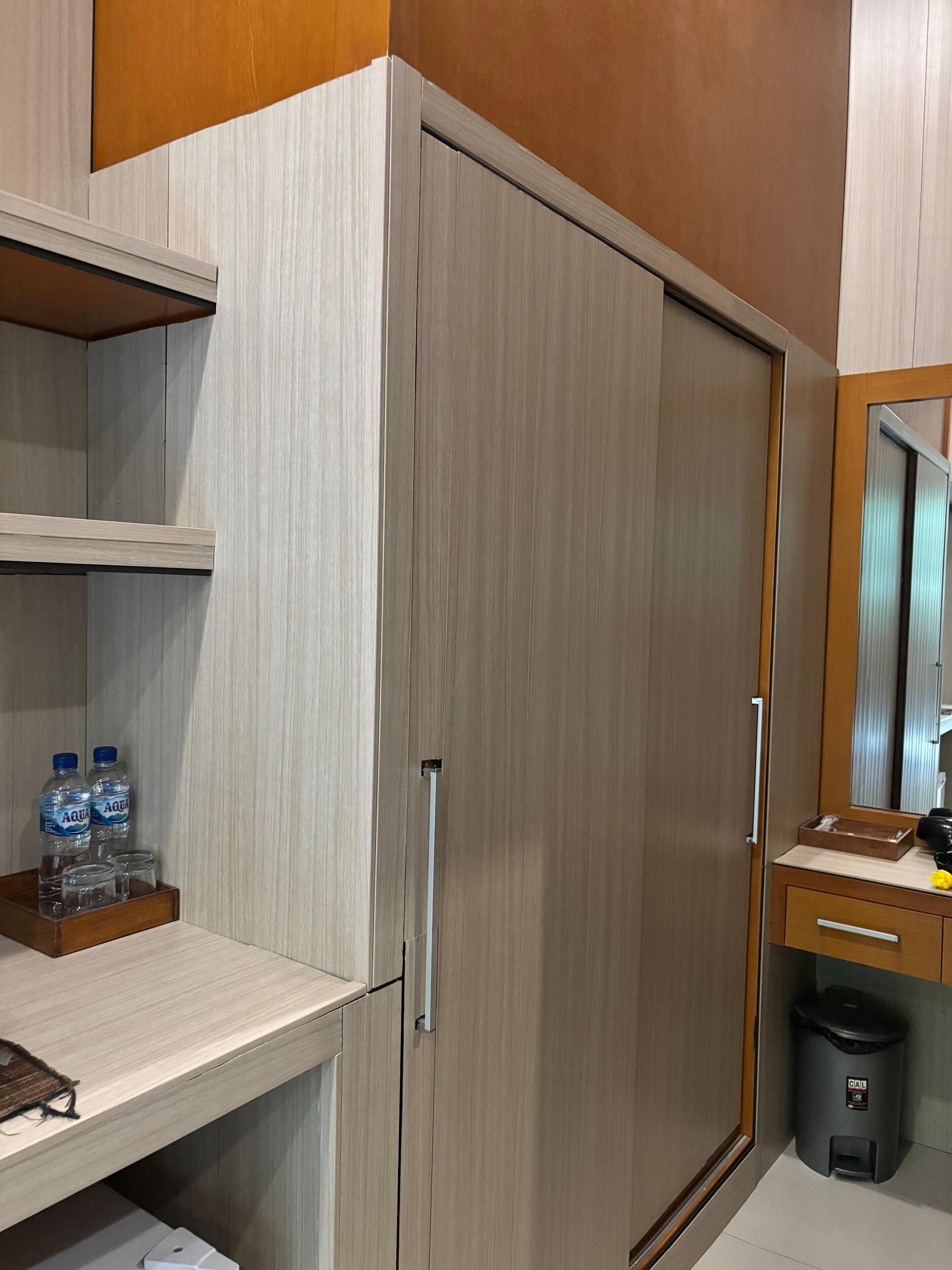 A hotel room with a wardrobe, a vanity with a mirror, a trash bin, and a shelf with water bottles and glasses.