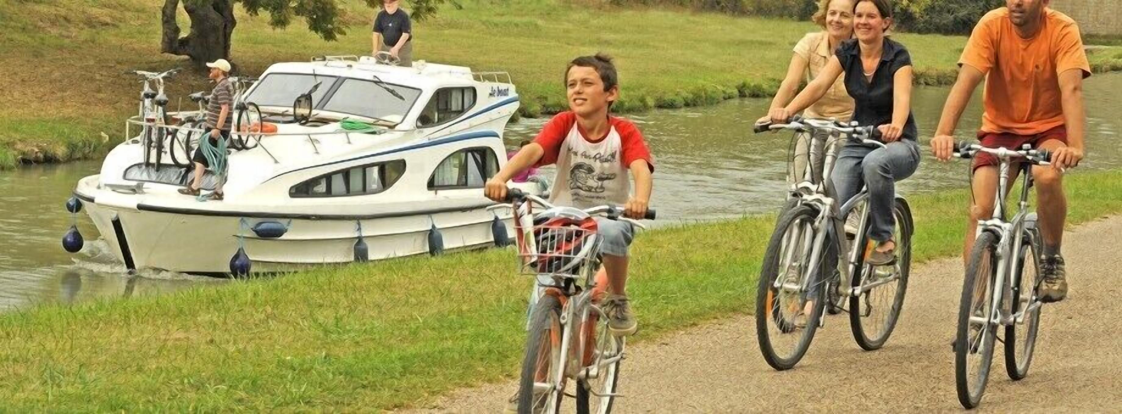 Een groep mensen die op fietsen rijdt langs een pad naast een rivier, met een boot afgemeerd in de buurt.