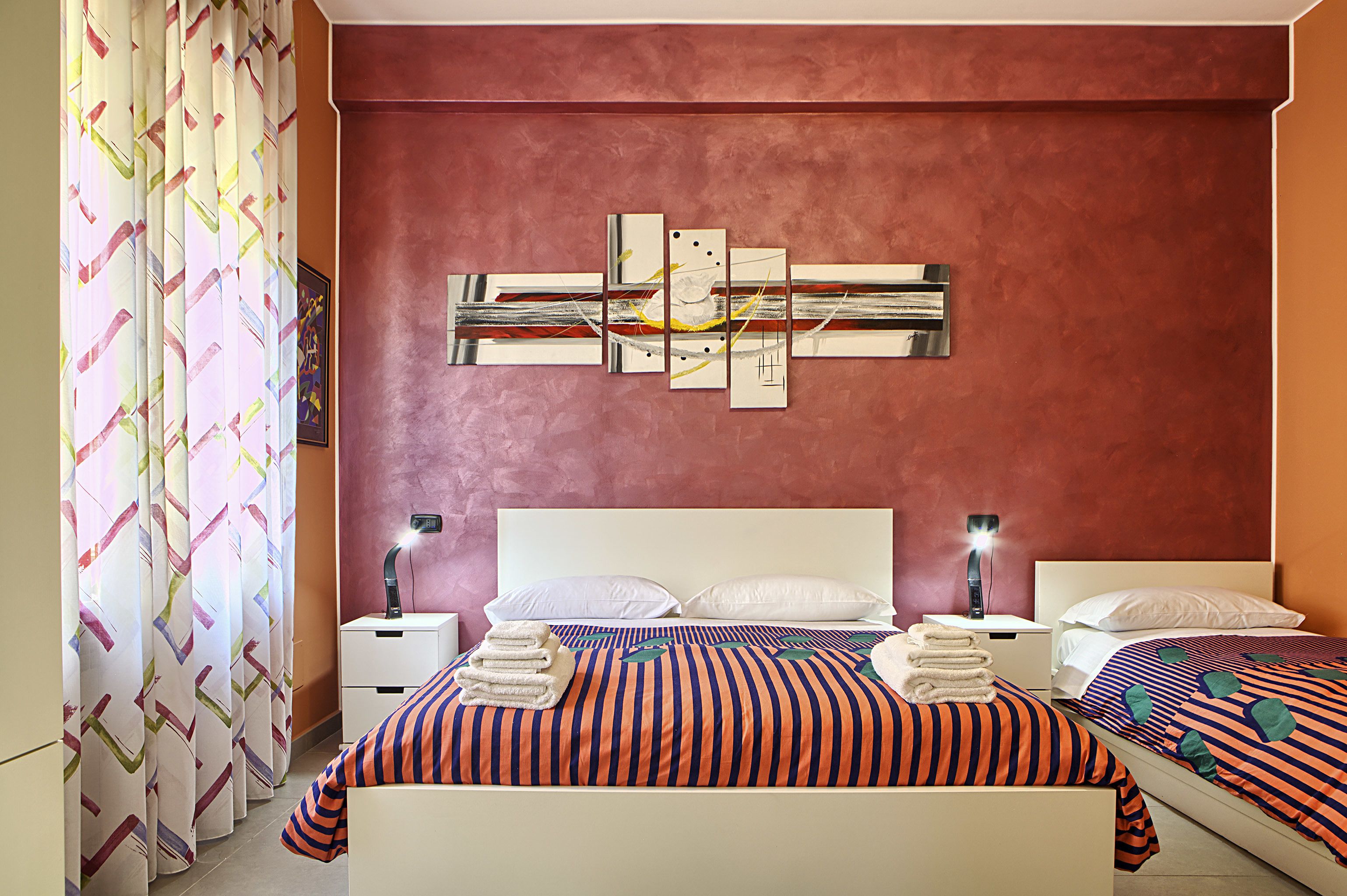 Quarto de hotel com uma cama grande, duas mesas de cabeceira com luminárias, uma obra de arte na parede e uma cortina estampada.