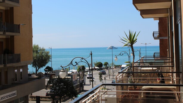 A balcony with a view of the sea, a restaurant with outdoor seating, and a building with a sign that reads "Assicurazioni". - La Collina dei Ciliegi (Salerno)
