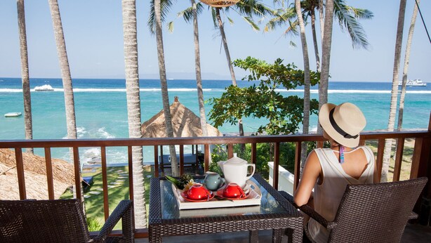 A balcony with wicker furniture, a table with red cups and a white teapot, and a view of the ocean with palm trees. - Anjani Villa (Manggis)