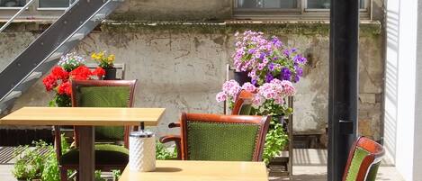 Un área de estar al aire libre con mesas y sillas de madera, rodeada de macetas con plantas y flores.