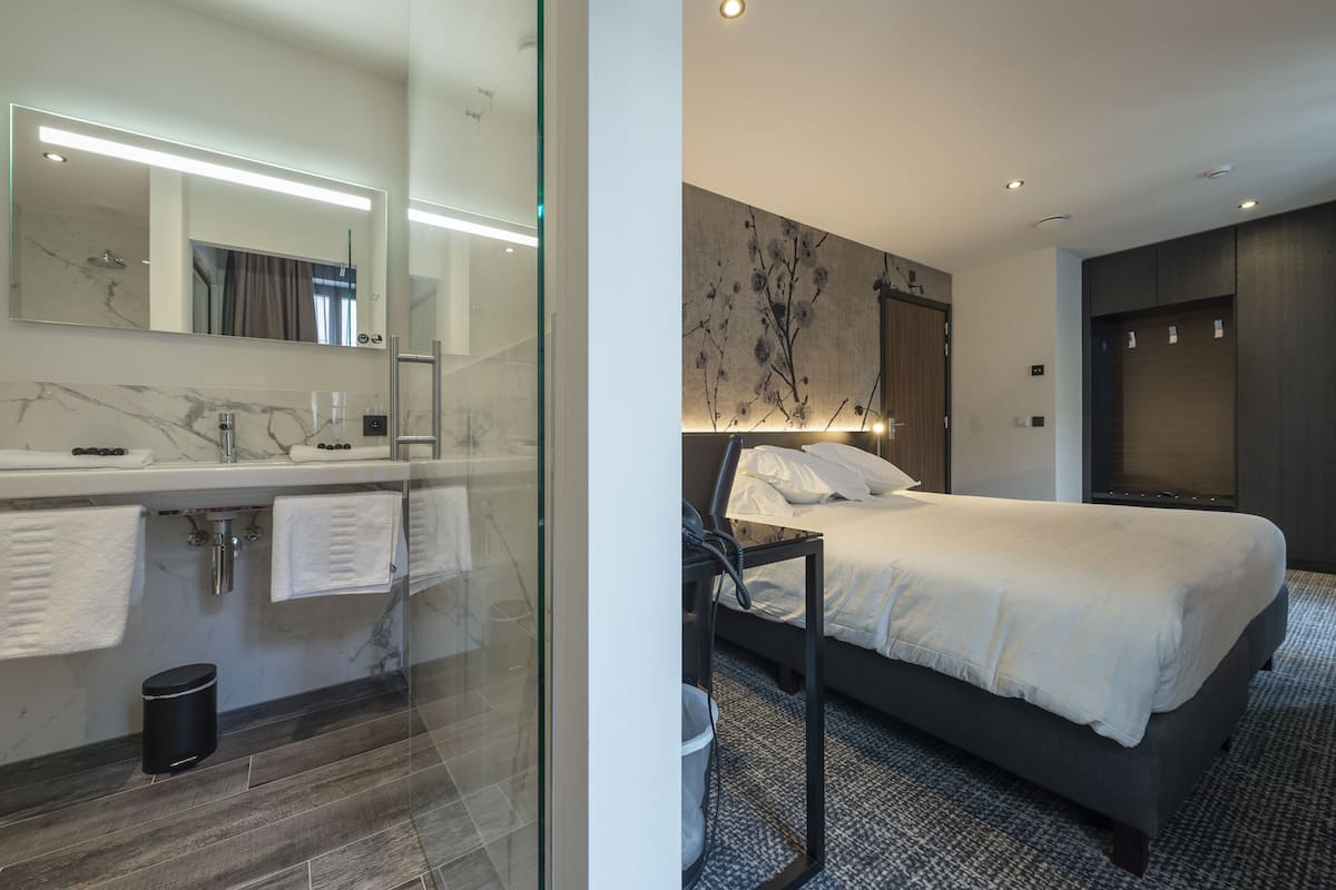 A modern hotel room with a glass-enclosed shower, a large bed with a headboard, a bedside table, and a wall-mounted light fixture.