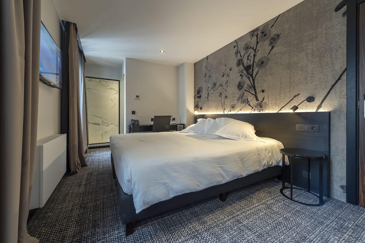 A modern hotel room with a large bed, a small table, a wall-mounted TV, and a large abstract painting.