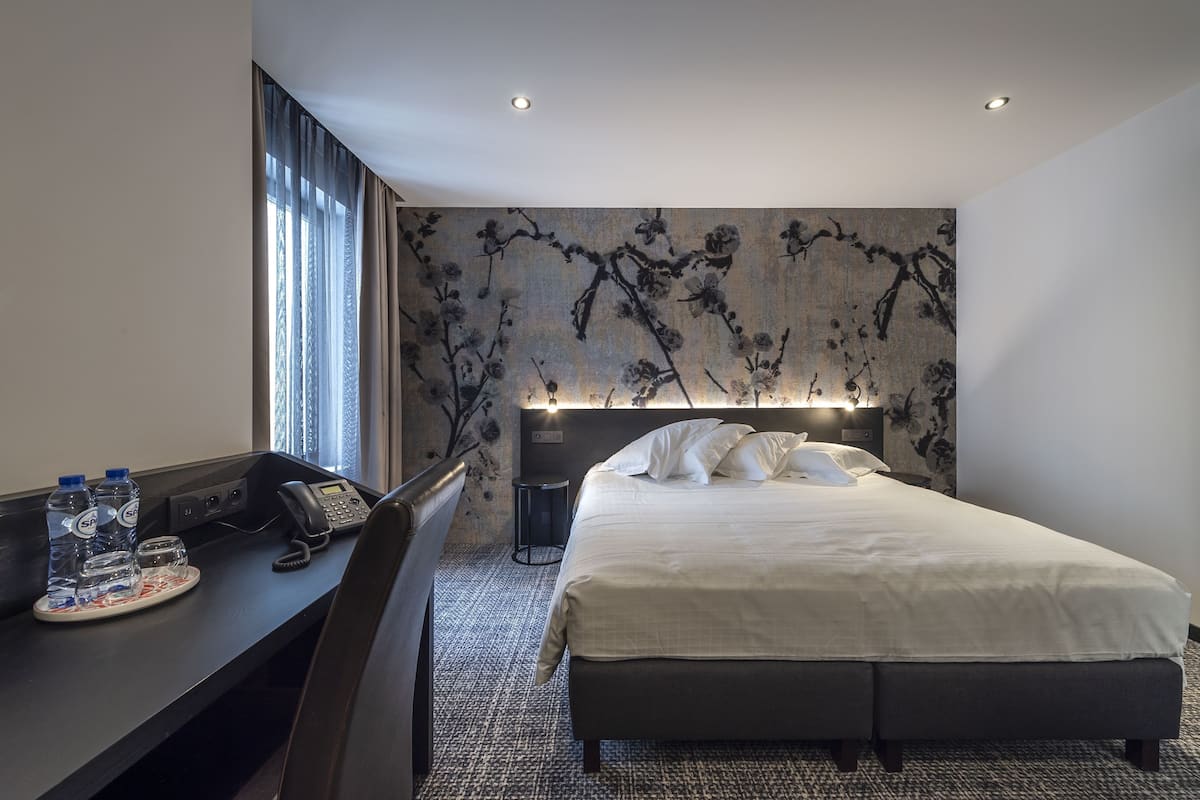 A hotel room with a bed, a desk with a phone, water bottles, and a wall with a floral design.