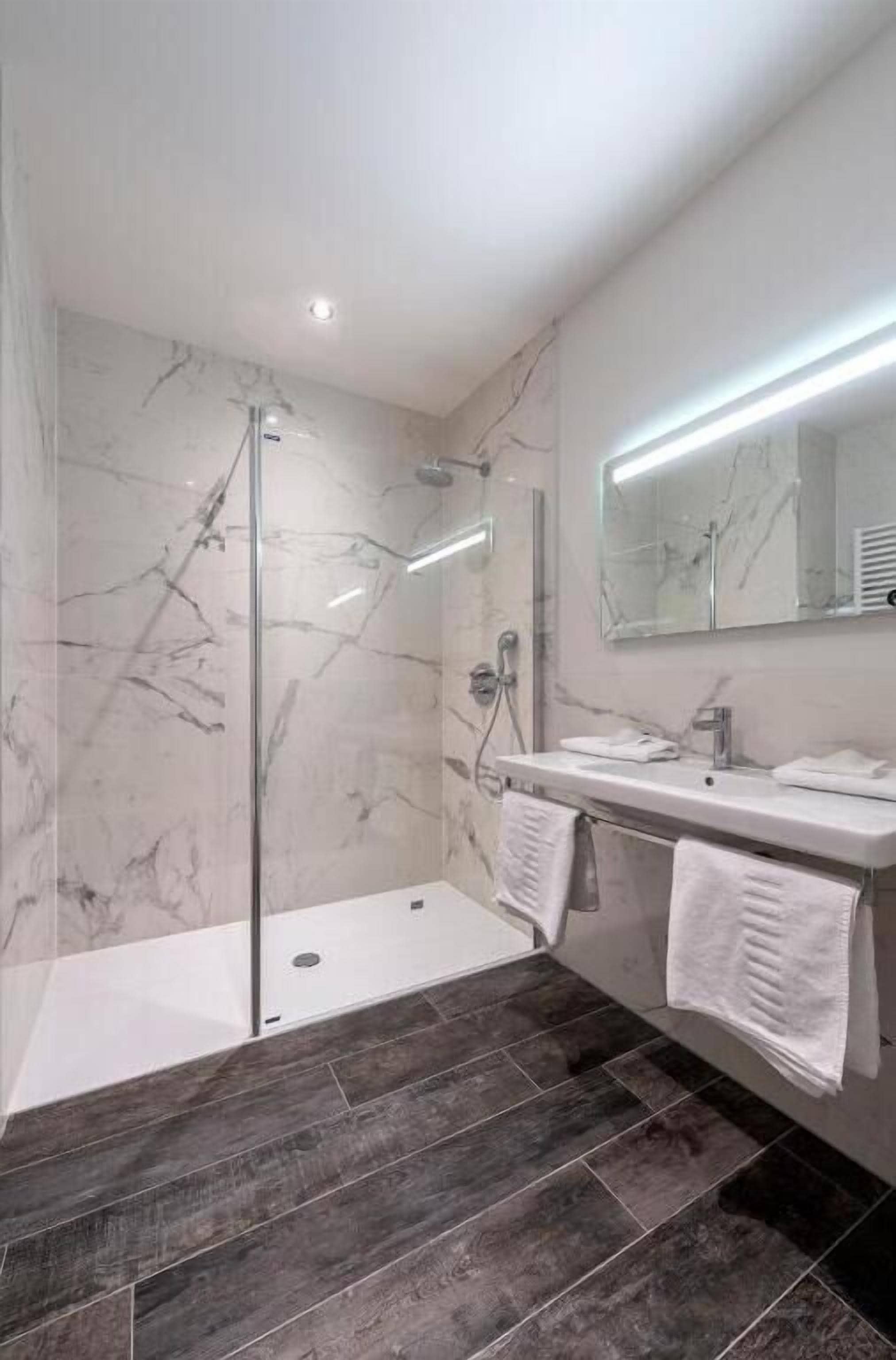 Un bagno moderno con cabina doccia in vetro, pareti in marmo, lavabo bianco e pavimento piastrellato in tonalità scura.