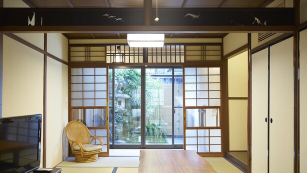 Et traditionelt japansk rum med tatamimåtter, et lavt bord, et fjernsyn på et stativ og skydedøre, der fører til en have.